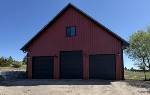 Red barn-style building with a steep roof, three large black garage doors, a single upper window, and outdoor lanterns, set on a dirt driveway with a tree and grassy area nearby.