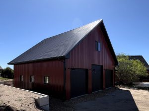 A red metal barn with a black corrugated roof, two large garage doors, and several windows under a clear blue sky.