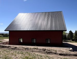 A red barn with a steep black metal roof sits on a dirt lot under a clear blue sky, with green grass and trees in the background.