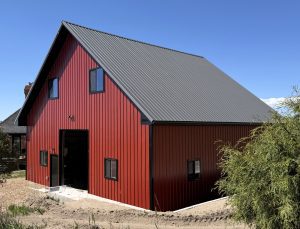 A modern red barn with a steep gray metal roof, black-trimmed windows, and a concrete entryway, set under a clear blue sky.
