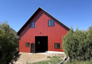 A red barn with a steep roof and black trim, featuring four windows and an open central door, stands among green bushes under a clear blue sky.