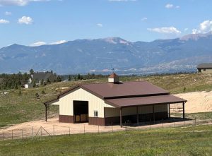 A tan and brown barn with a covered side area stands on a grassy hill, with mountains and a few houses visible in the background under a partly cloudy sky.