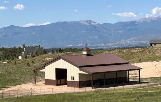 A tan and brown barn with a covered side area stands on a grassy hill, with mountains and a few houses visible in the background under a partly cloudy sky.