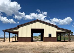 A beige and brown metal barn with a central open bay and two covered side wings, set on a dirt lot under a blue sky with scattered clouds.
