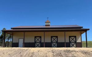 A tan and brown barn with four doors, a side entry door, and a small cupola on the roof, set against a clear blue sky.