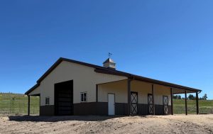A tan and brown barn with a weather vane on the roof, three doors, and windows, set on dry ground under a clear blue sky.