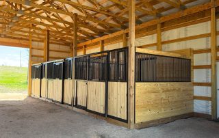 Four wooden and metal horse stalls inside a partially enclosed barn with a dirt floor and exposed wooden beams.