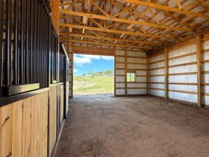 Interior of a wooden barn with exposed beams, one horse stall on the left, and an open doorway leading to a grassy outdoor area under a blue sky.