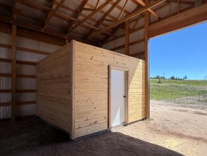 A small wooden room with a white door is built inside a larger open-walled barn structure, with sunlight and grassy field visible outside.