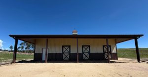 A metal barn with three doors stands on a sandy lot under a clear blue sky, with open land and trees in the background.