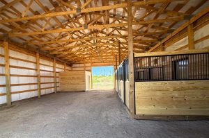 Interior of a wooden horse stable with empty stalls on the right and an open doorway at the far end letting in natural light.