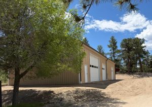 A beige metal building with three large white garage doors stands on a dirt lot, surrounded by trees under a partly cloudy sky.