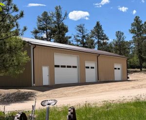 A large beige metal garage building with three white overhead doors and a single entry door stands beside a dirt driveway, surrounded by trees under a blue sky.