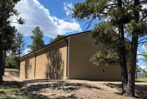A large tan metal building sits on bare ground, surrounded by tall pine trees under a partly cloudy blue sky.