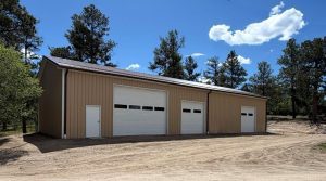 A large beige metal garage building with three white roll-up doors and one side entrance door, surrounded by trees and a dirt driveway under a blue sky with clouds.