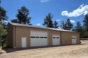 A large beige metal building with three white garage doors and one entry door sits on a dirt lot surrounded by pine trees under a partly cloudy sky.