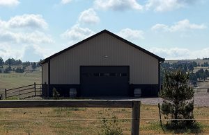 A beige metal barn with a large dark garage door sits on a rural property, surrounded by fencing, trees, and grassy fields under a partly cloudy sky.