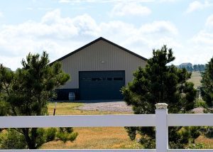 A beige metal barn with a large closed garage door is partially obscured by pine trees and a white fence in the foreground.