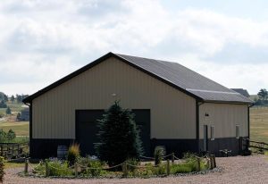 A beige metal barn with two large garage doors stands on gravel, surrounded by a wooden fence, small trees, and pastureland under a partly cloudy sky.