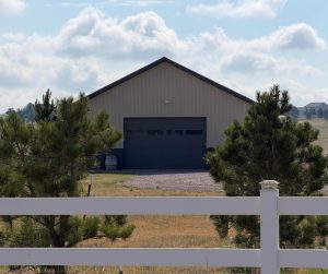 A beige metal garage with a large dark door stands behind a white fence and two pine trees on a rural property under a partly cloudy sky.