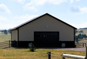 A tan and black metal barn with a closed garage door sits in a grassy, rural landscape under a partly cloudy sky.