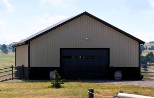 A tan metal barn with a large dark garage door sits in a grassy field, surrounded by a wooden fence and a few small trees.