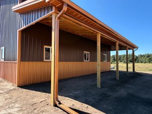 A metal building with vertical siding features a covered porch supported by wooden posts, situated on bare soil under a clear blue sky.