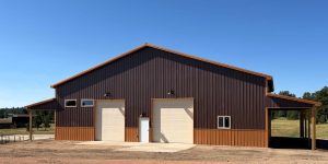 Large brown metal barn with three white doors, small windows, and a clear blue sky. The barn stands on a concrete slab surrounded by open land and some trees.