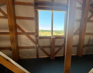 Unfinished loft area with exposed wooden framing and insulation. A window reveals a view of trees under a clear blue sky.