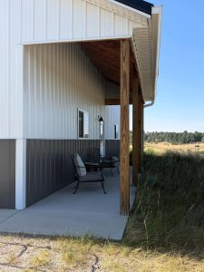 A covered porch with two metal chairs and a bench sits beside a metal-sided building, overlooking grassland under a clear blue sky.