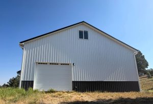 A large white barn with a black base, a garage door, and a small window, standing on dry grass under a clear blue sky.