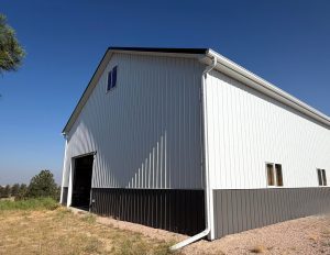 A white metal barn with a black lower section stands on a gravel lot under a clear blue sky. One large door is partially open.