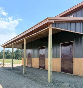 A metal-sided building with a covered porch, three brown doors, and wooden posts, set on a dirt lot with trees in the background.