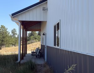 A covered porch with two benches sits beside a modern white metal building, surrounded by dry grass and trees under a clear sky.