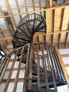 A metal spiral staircase leads up to a loft in a building with exposed wooden beams and insulation on the ceiling and walls.