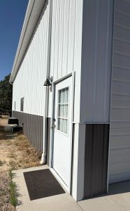 White and gray metal building with a white door, outdoor light fixture above, black doormat, and window panel on the door; sidewalk and gravel visible.