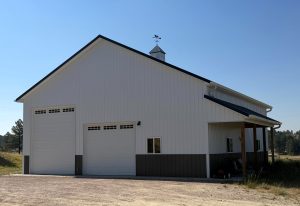 A large white metal building with two garage doors, a small window, covered side area, and cupola, situated on gravel with grass and trees in the background under a clear sky.