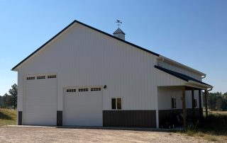 Colorado pole barn in a cleared rural setting with gravel access and pine trees positioned beyond the building site