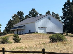 A white barn with a metal roof sits on a grassy hill, surrounded by trees and a wooden fence under a clear blue sky.