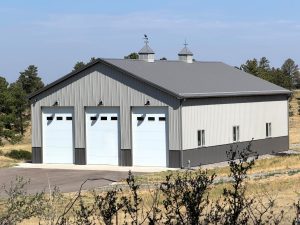 A metal building with three large garage doors, gray siding, and two cupolas on the roof, set in a rural area with trees and grass.