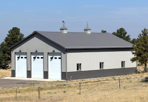 Gray metal building with three large garage doors and small windows on the side, set in a grassy area with scattered trees and a clear sky in the background.