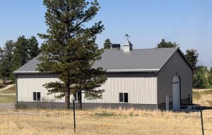 A gray metal barn with a pitched roof and cupola sits in a grassy field near tall trees, with a large arched door and several windows visible.