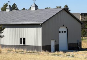 Large gray metal barn with a peaked roof, two cupolas, one window, one large garage door, and a small entry door, surrounded by dry grass and trees.