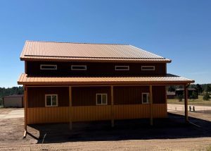 A large brown and yellow metal barn with a sloped roof, four small upper windows, and a covered porch, set in a rural area under a clear blue sky.