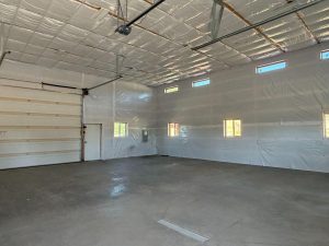 Large, empty garage with concrete floor, insulated walls and ceiling, multiple windows, fluorescent lights, and a closed overhead door.