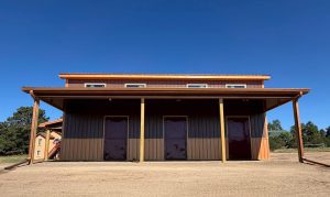 A brown metal barn with three closed doors and a covered porch stands on a dirt lot under a clear blue sky.