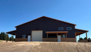 A large, dark brown metal building with a white garage door and two white entry doors, situated on a dirt lot under a clear blue sky.