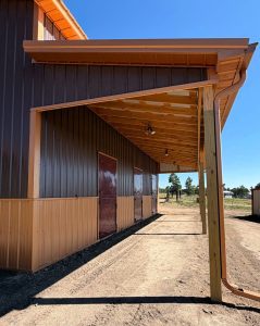 A brown and tan metal building with a covered porch supported by wooden posts, casting shadows on the dirt ground under a clear blue sky.