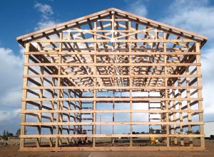 Wooden framework of a large, unfinished barn or warehouse, viewed from the front, under a partly cloudy sky.