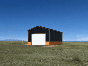 A black and orange metal barn with a large white door stands alone on a grassy plain under a clear blue sky.